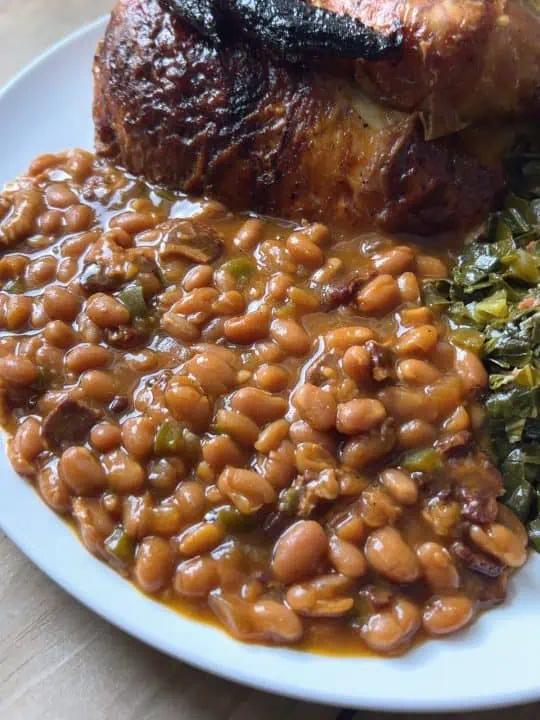 Baked beans on a white plate with roasted chicken and collard greens.