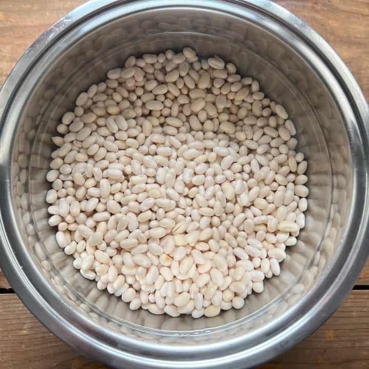 Dry navy beans soaking in water in a stainless steel bowl on a wood table.