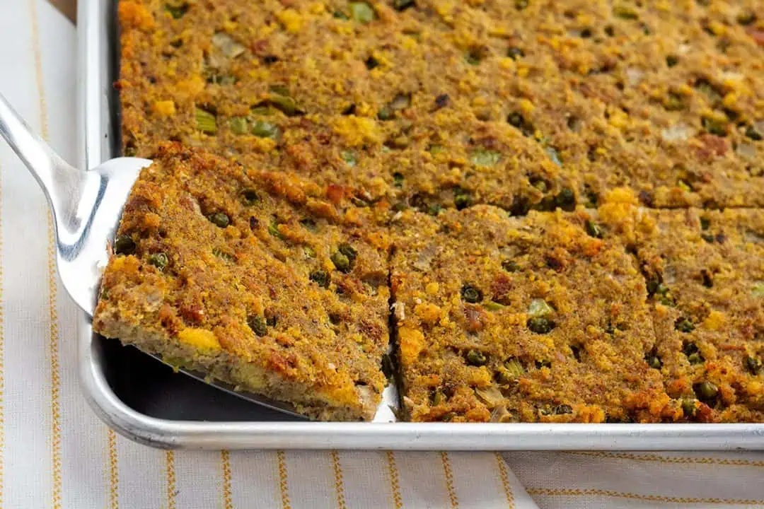 Lifting a slice of Southern cornbread dressing on a spatula from a half-sheet pan.