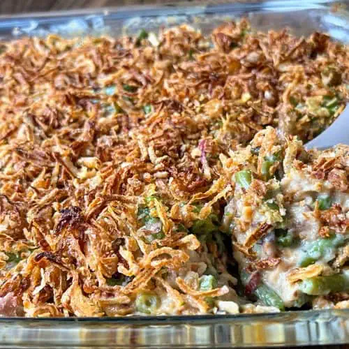 A casserole dish filled with green beans topped with crispy onions, being scooped with a serving spoon.