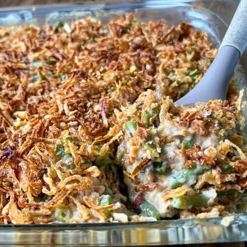 A serving spoon scooping a portion of vegan green bean casserole topped with crispy fried onions from a glass baking dish.