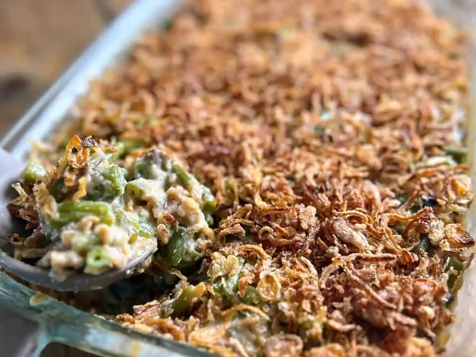 A casserole dish filled with green beans topped with crispy onions, being scooped with a serving spoon.