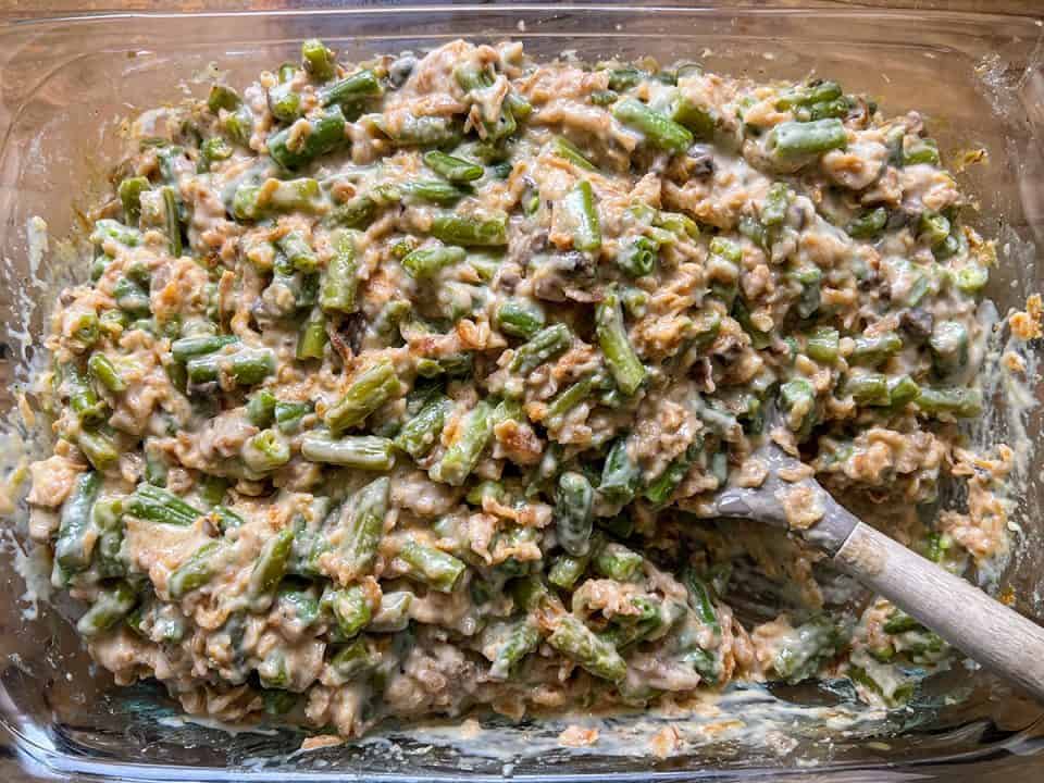 A casserole dish containing a mix of green beans, creamy soup, and crispy onions being stirred.