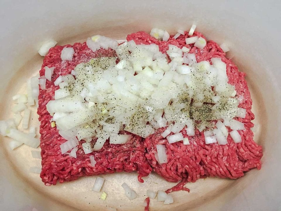 A Dutch oven with uncooked ground beef topped with diced onions, salt, and pepper.