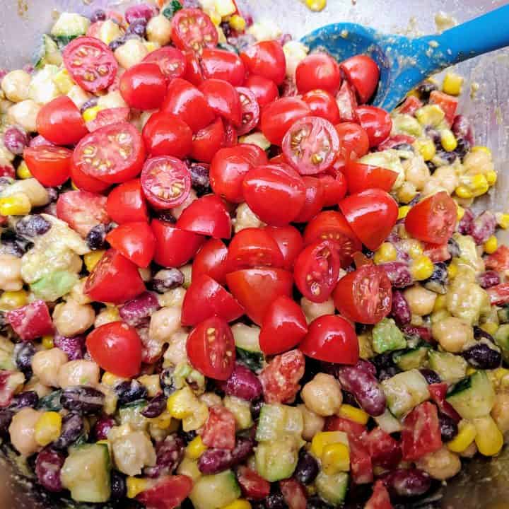 Mixing canned bean salad topped with grape tomatoes in large bowl with teal spoon.