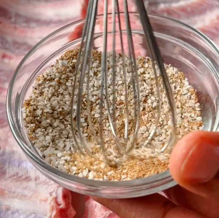 Holding a small glass bowl and combining spices with a whisk, with ribs in the background.