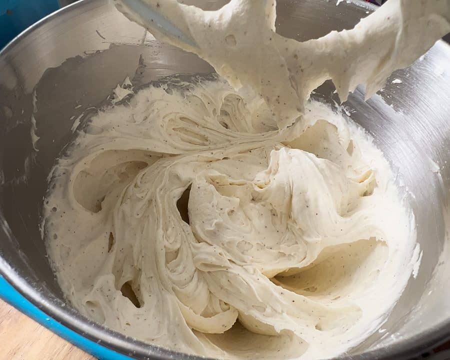 Whipped brown butter cream cheese frosting in a stand mixer, smooth and fluffy with visible specks.