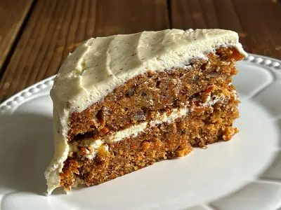 A close-up slice of two-layer hummingbird carrot cake on a white plate, showing moist texture with visible flecks of carrot, pineapple, and pecans, topped with brown butter cream cheese frosting.