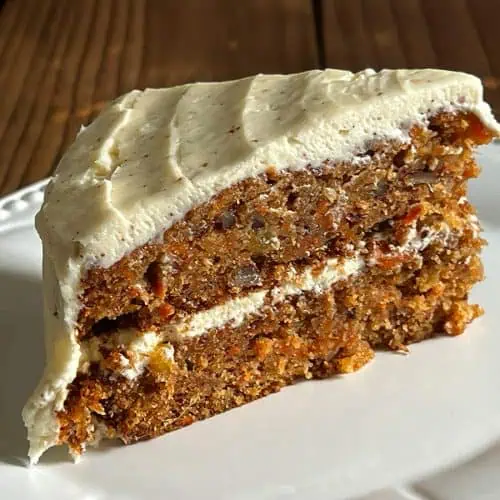 A close-up slice of two-layer hummingbird carrot cake on a white plate, showing moist texture with visible flecks of carrot, pineapple, and pecans, topped with brown butter cream cheese frosting.