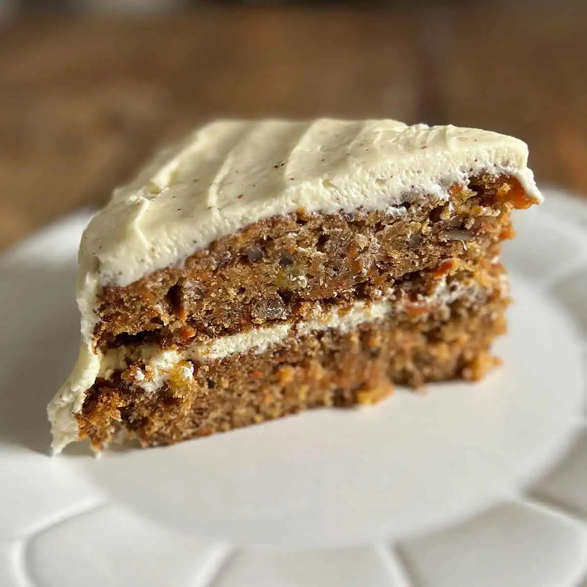 A single slice of hummingbird carrot cake with cream cheese frosting, served on a white plate with a wooden table in the background, square format for social sharing.