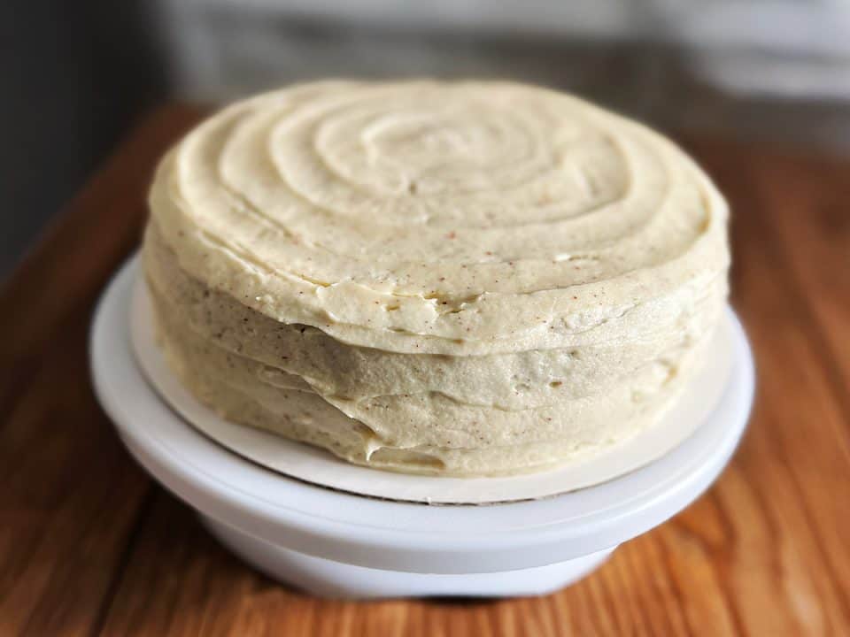 A fully frosted hummingbird carrot cake with smooth, swirled brown butter cream cheese frosting, displayed on a white cake stand over a wood surface.