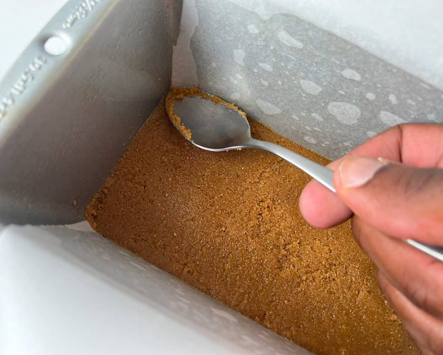 Hand pressing graham cracker crust into a 12x4 baking pan using a spoon, lined with parchment paper