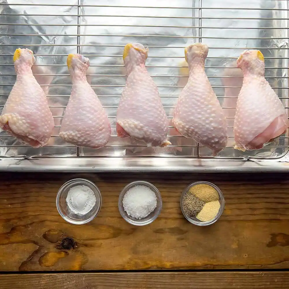 Five raw chicken drumsticks on a rack with bowls of baking powder, kosher salt, and spices underneath--dry brine setup.