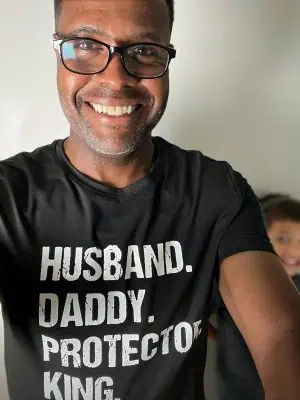 Gary White smiling in a black t-shirt that reads “Husband. Daddy. Protector. King.” with his son smiling in the background.