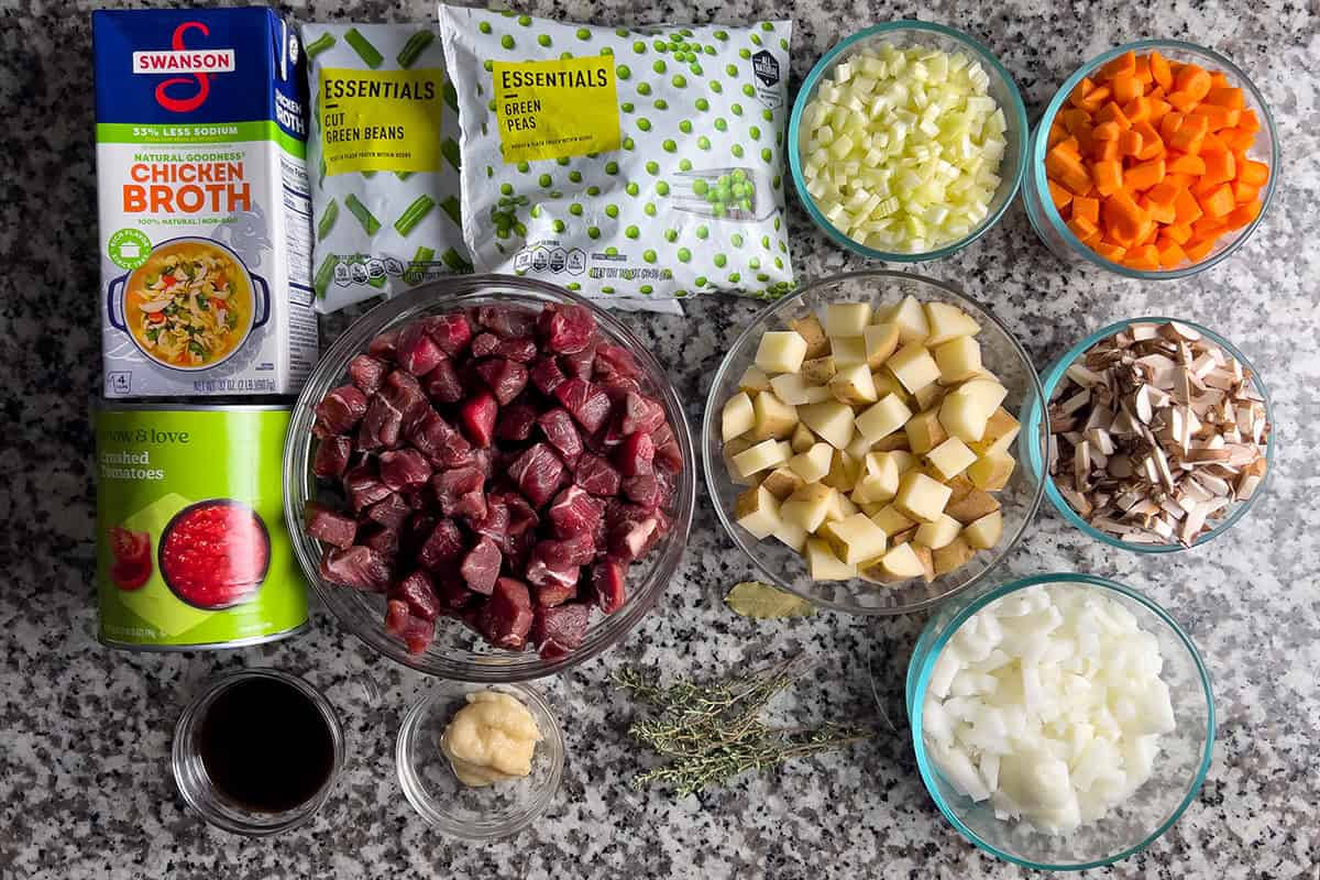 Ingredients for Instant Pot vegetable beef soup arranged on countertop, including beef, potatoes, carrots, celery, onions, mushrooms, green beans, peas, broth, and crushed tomatoes