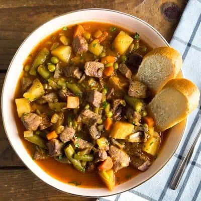 Bowl of vegetable beef soup with potatoes, carrots, green beans, and bread slices on the side