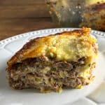 Slice of pot roast lasagna on a white plate with the pan in the background, showing stacked noodles, shredded roast, and melted cheese.