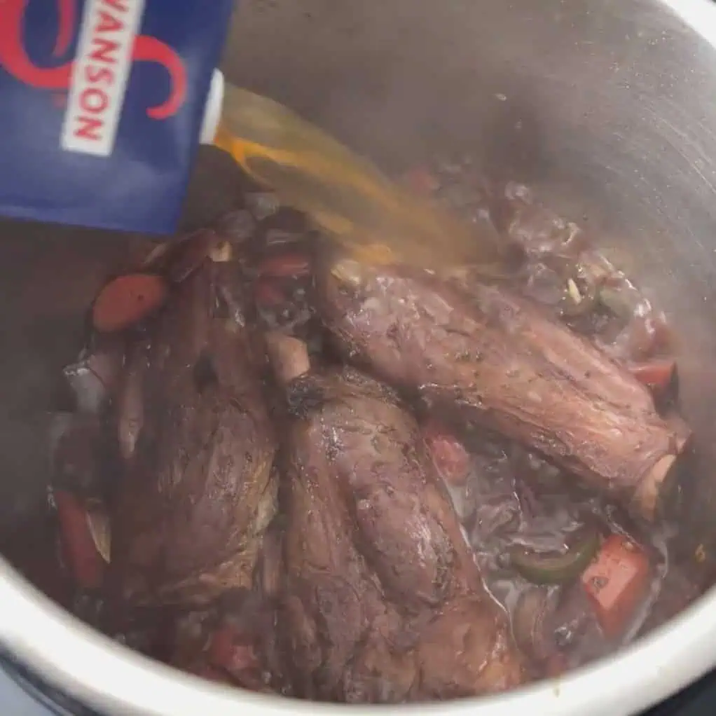 Broth being poured into the Instant Pot over lamb shanks and vegetables to create the braising liquid.