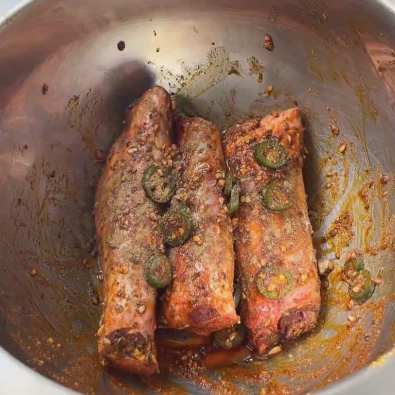 Lamb shanks marinating with garlic, spices, and sliced green chile in a metal bowl before cooking.