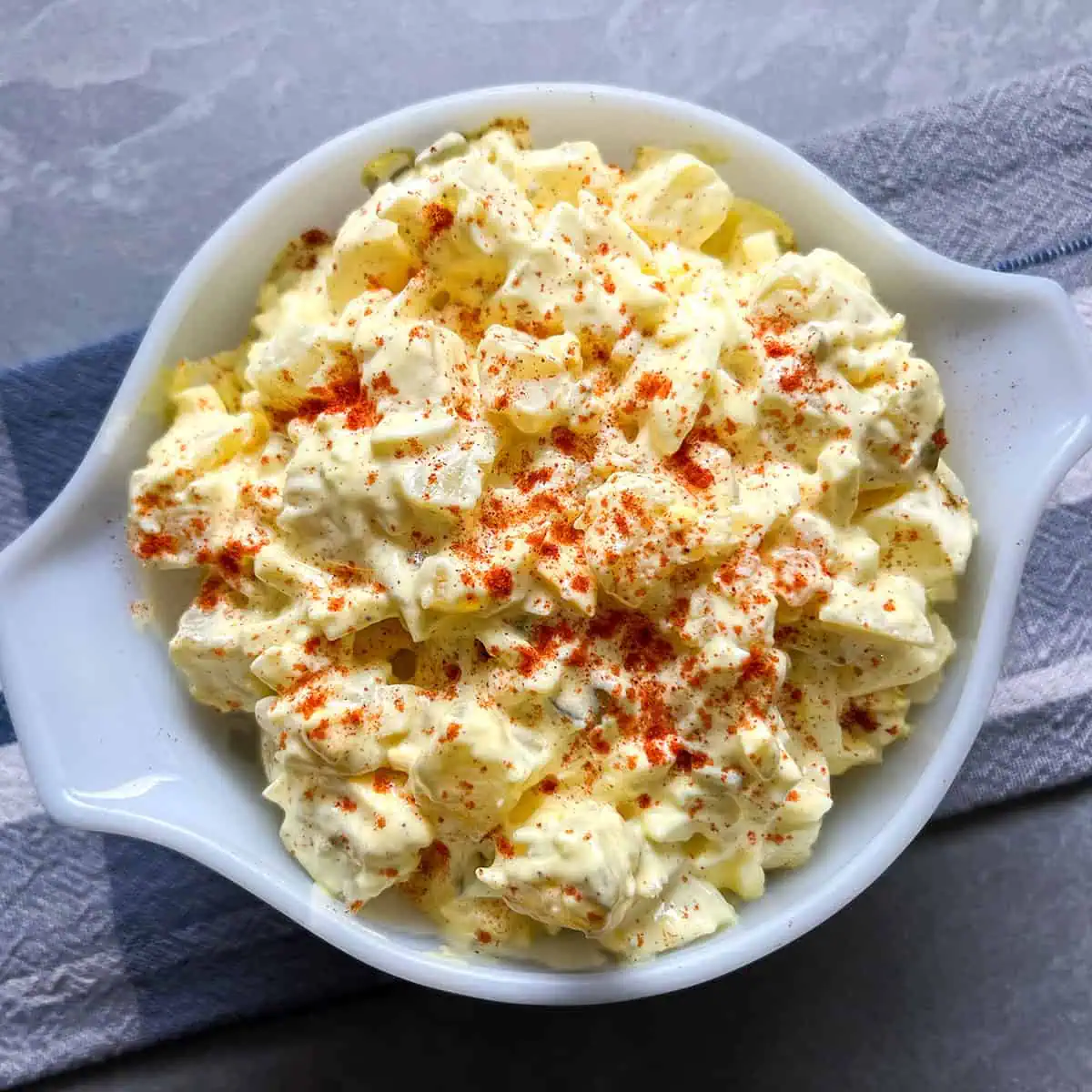 Close overhead view of creamy deviled egg-style potato salad topped with paprika in a white dish.