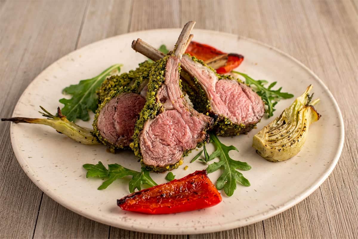 Sliced mint-crusted rack of lamb showing a pink center on a plate with roasted fennel and red bell pepper