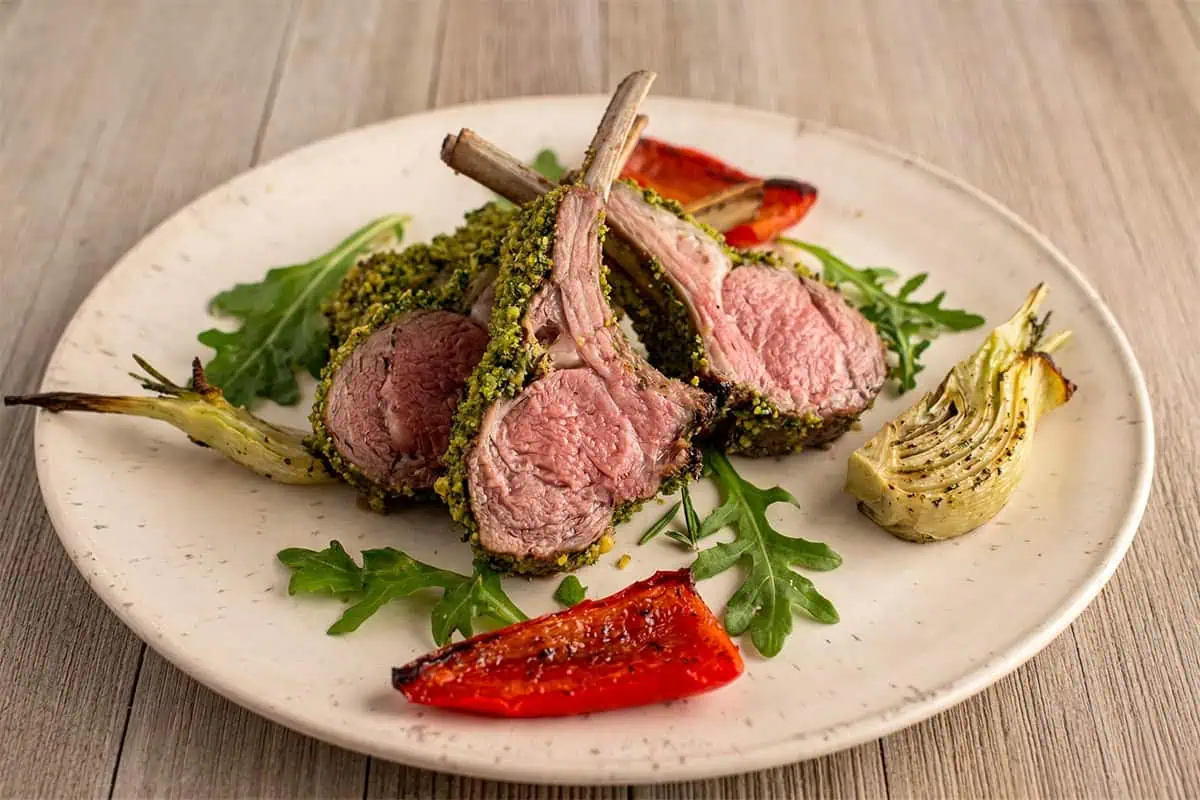 Sliced mint-crusted rack of lamb showing a pink center on a plate with roasted fennel and red bell pepper