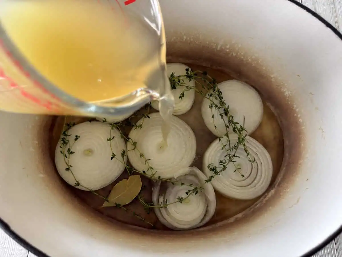 Chicken broth being poured into a Dutch oven with sliced onion, thyme, and bay leaf for oven corned beef.