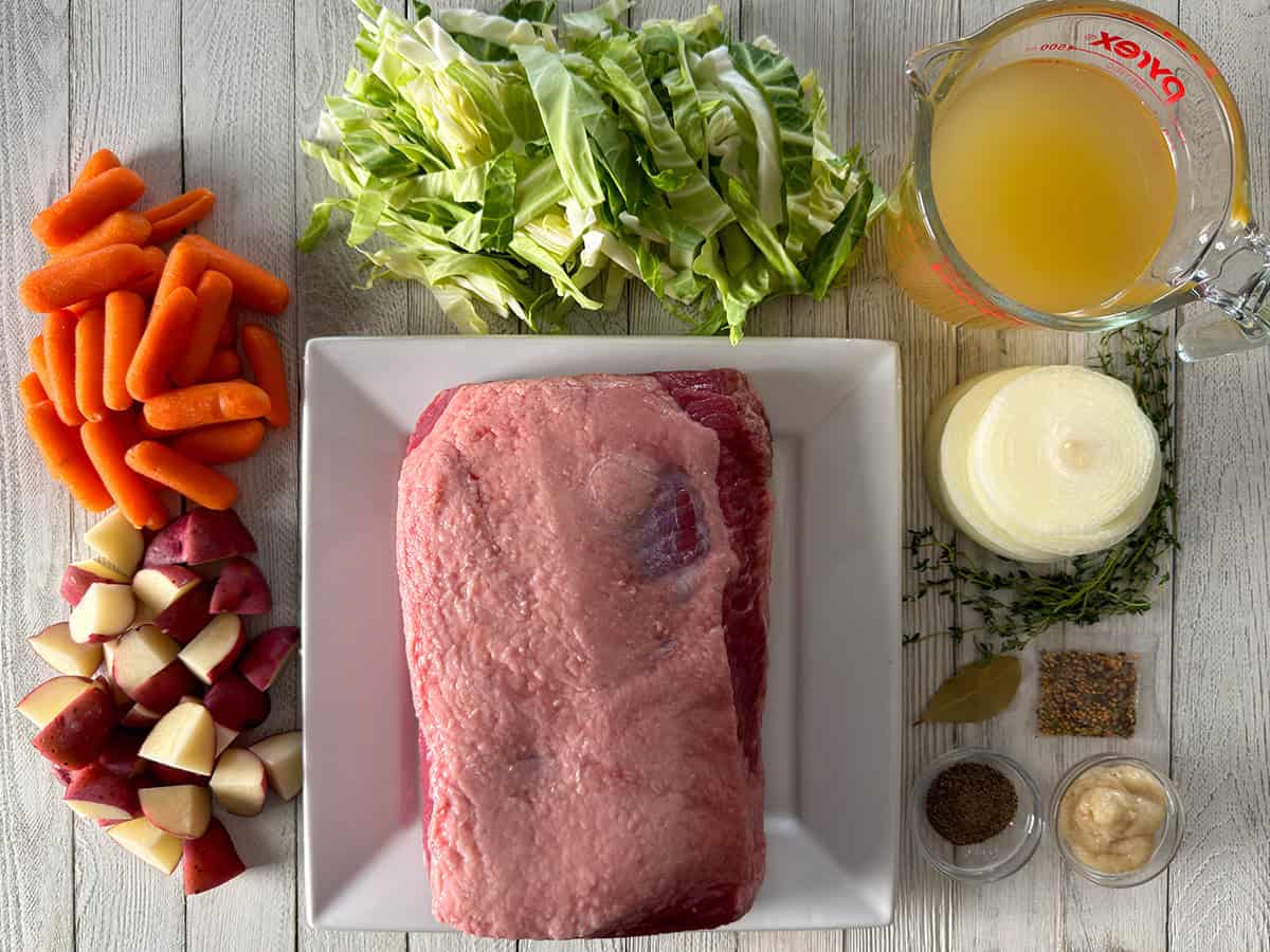 Ingredients for oven corned beef including corned beef brisket, chicken broth, cabbage, carrots, red potatoes, onion, thyme, bay leaf, garlic paste, black pepper, and the spice packet.