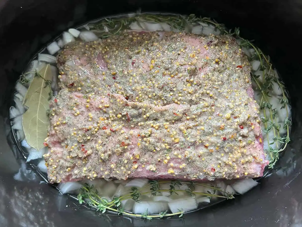 Corned beef coated with garlic and spice paste placed over onions, thyme, and bay leaf in a slow cooker.