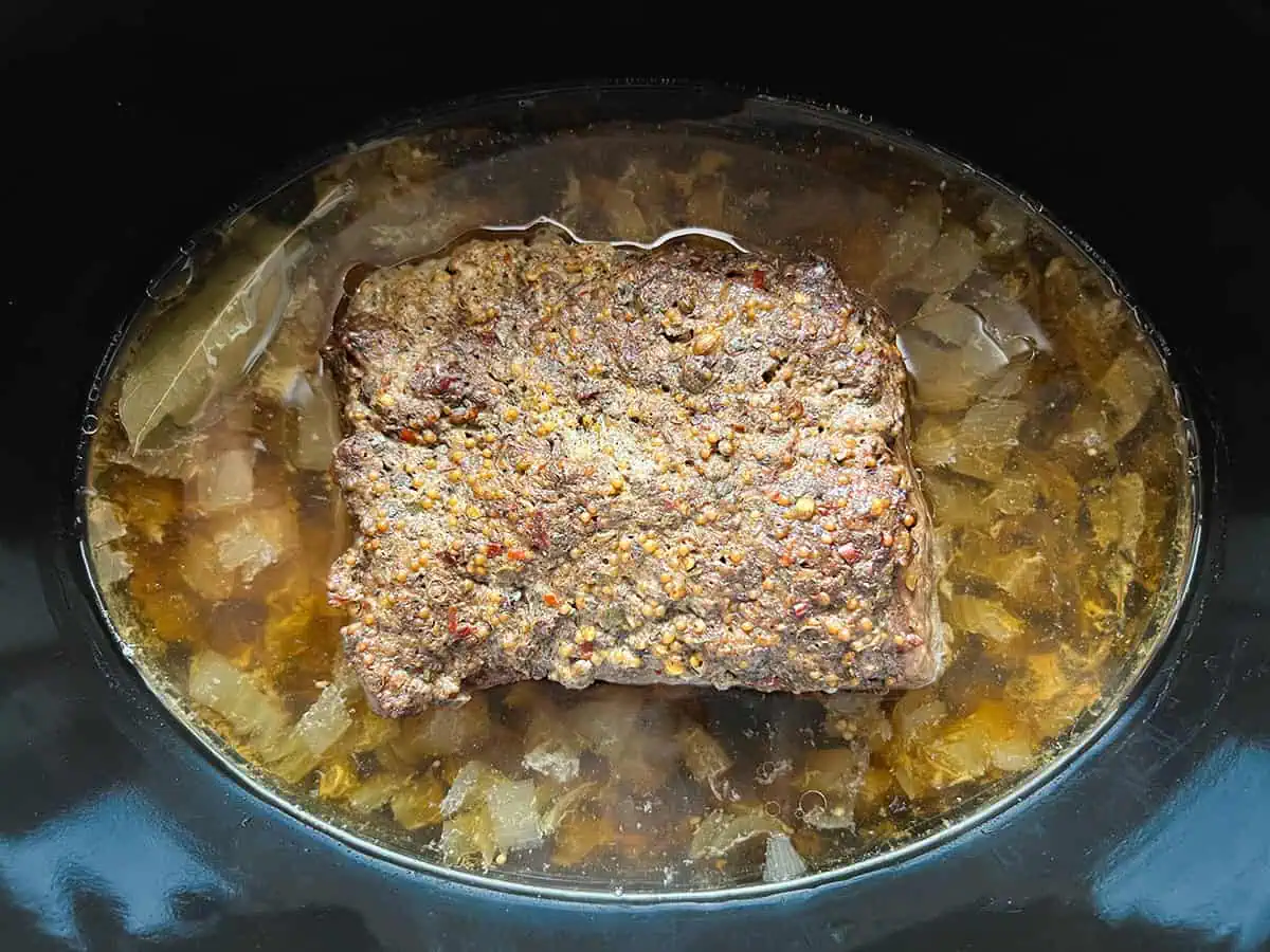 Corned beef with garlic and spice topping cooking in broth with onions and bay leaves in a slow cooker.