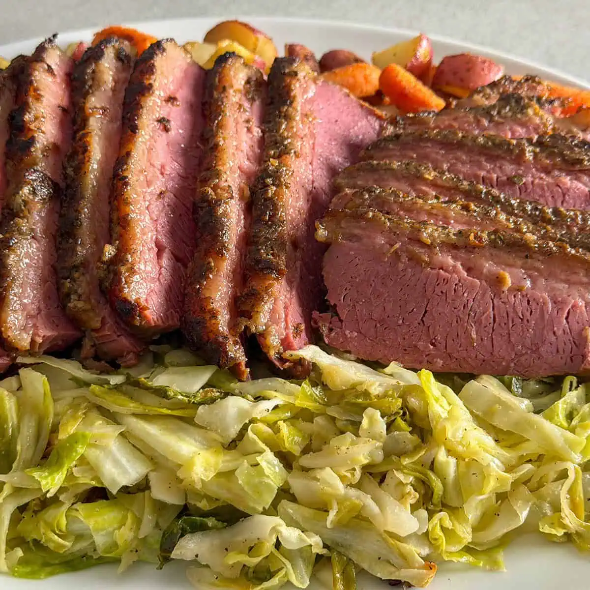 Sliced slow cooker corned beef with crispy garlic top served with roasted red potatoes, carrots, and cabbage on a white platter.