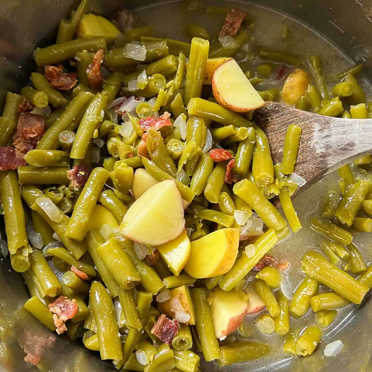 Cooked southern green beans with potatoes and bacon in a pot with a wooden spoon.