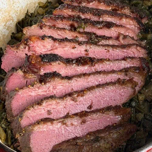 Sliced corned beef with a dark crust served over greens in a Dutch oven.