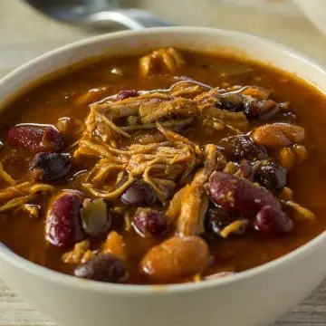 A close-up of a bowl of chili with shredded chicken, kidney beans, and black beans in a rich tomato sauce. A spoon is visible in the blurred background.