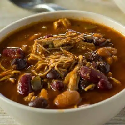 A close-up of a bowl of chili with shredded chicken, kidney beans, and black beans in a rich tomato sauce. A spoon is visible in the blurred background.