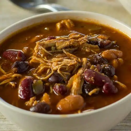 A close-up of a bowl of chili with shredded chicken, kidney beans, and black beans in a rich tomato sauce. A spoon is visible in the blurred background.