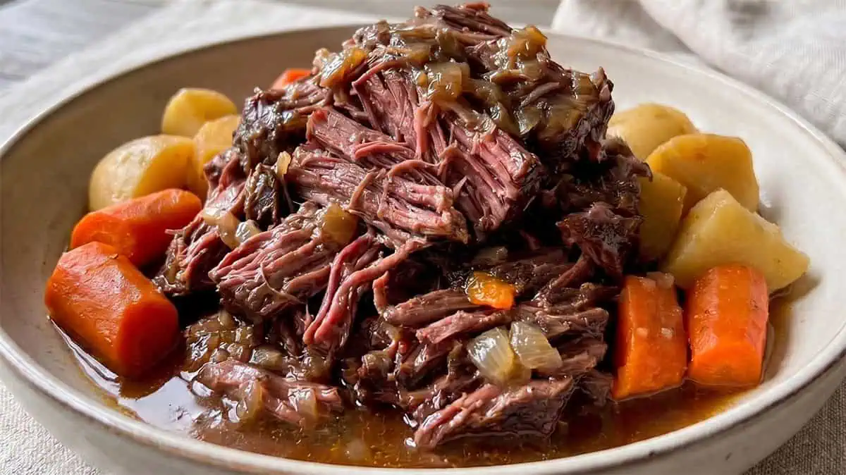 A plate of pot roast with shredded beef, surrounded by chunks of carrots and potatoes, all in a rich brown gravy with chopped onions.