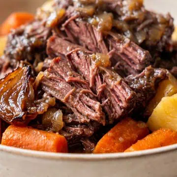Close-up of a tender pot roast topped with caramelized onions, served with chunks of roasted carrots and potatoes in a bowl. The meat appears juicy and flavorful.