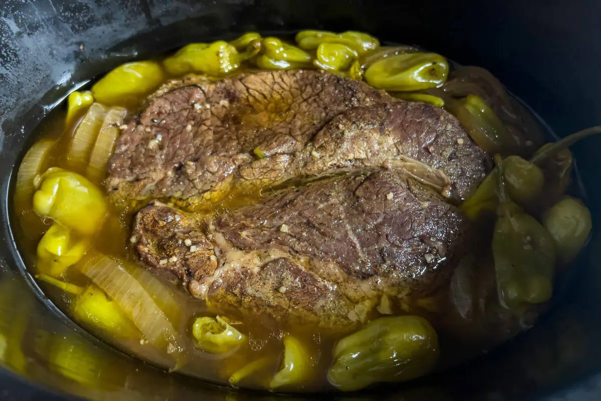 Chuck roast cooking in a slow cooker with pepperoncini peppers, sliced onions, and broth for Mississippi pot roast