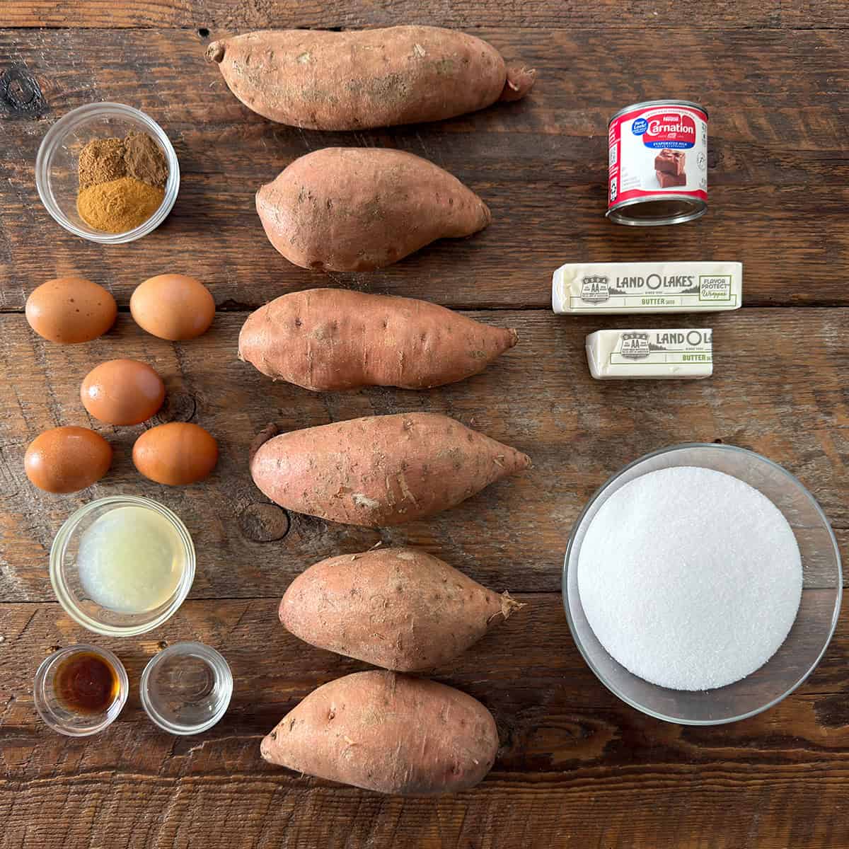 Sweet potato pie ingredients including sweet potatoes, eggs, butter, sugar, evaporated milk, lemon juice, extracts, and spices on a wooden table.