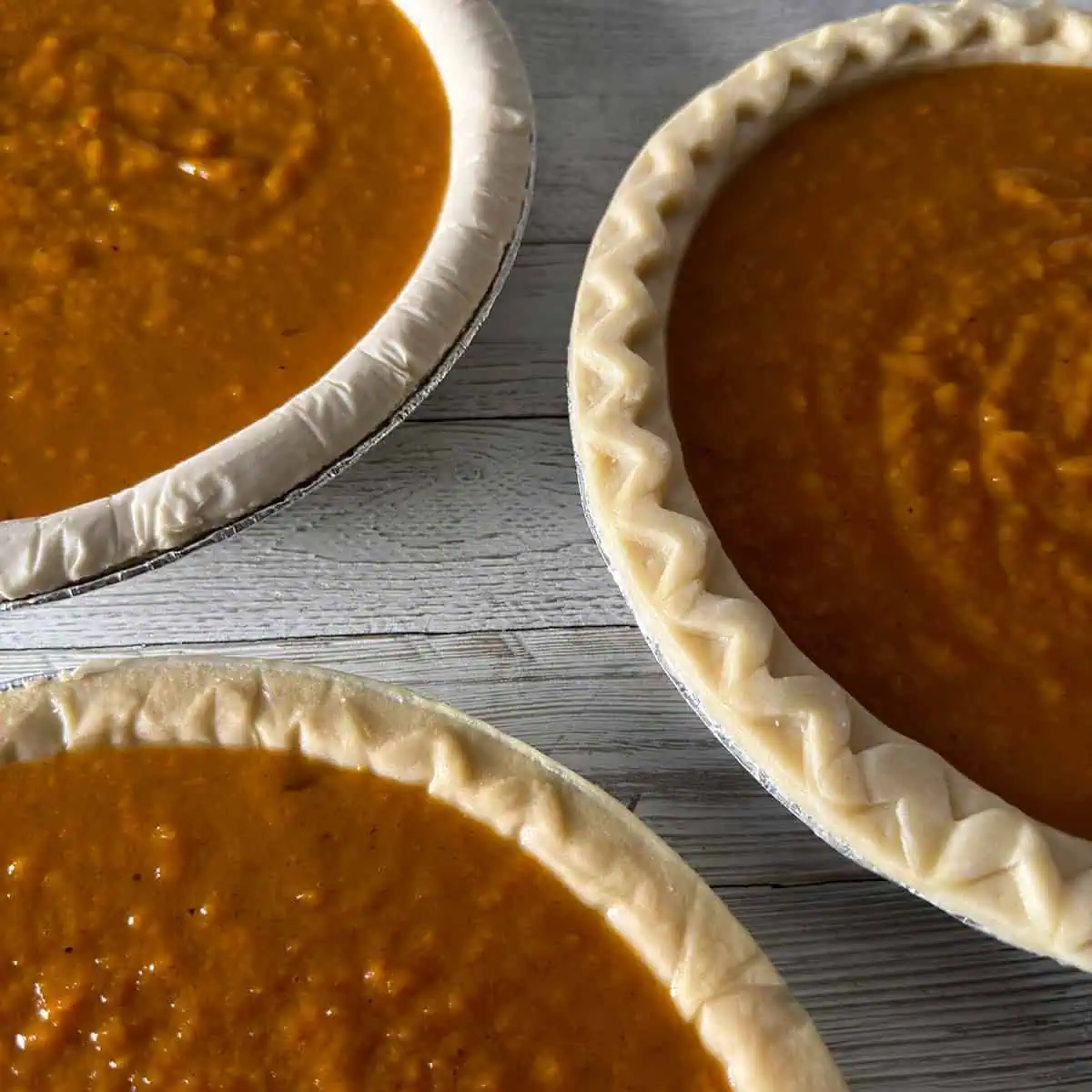 Three sweet potato pies filled in store-bought pie shells before baking on a light wood surface.