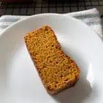 A slice of gluten-free pumpkin bread on a plate with additional loaves cooling on a rack in the background.